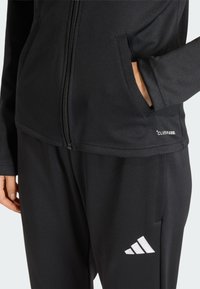 Μαύρο αθλητικό μπουφάν με φερμουάρ, πλαϊνές τσέπες και λογότυπο «CLIMACOOL». Συνδυάζεται με μαύρο παντελόνι που φέρει λευκό λογότυπο Adidas.