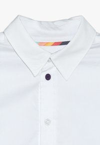 Valkoinen kauluspaita, jossa on violetti nappi, toinen valkoinen nappi ja värikäs etiketti sisäkauluksessa. Silkkinen, rapea kangastuntuma.