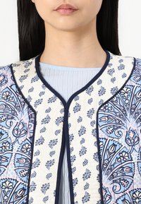 Chaleco acolchado con intrincados patrones florales en azul y rosa sobre un fondo crema, con ribete en azul marino y un acabado suave y texturizado.