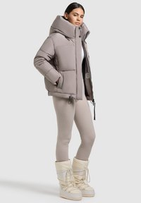 Giacca puffer di un beige chiaro con colletto alto, chiusura con zip e tasche laterali. Abbinata a stivali da neve color crema e leggins beige.