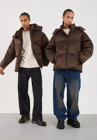 Giubbotti puffa marroni, con zip e dettagli del colletto a scatto. Abbinati a pantaloni in denim scuri e sbiaditi, e scarpe nere con lacci. Sfondo neutro.