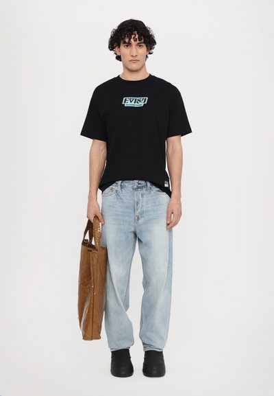 Melnas kokvilnas T-krekls ar tirkīza logotipu, gaiši zili džinsi, brūna rokassoma un melni slip-on apavi. Vienkārša, ikdieniška apģērbu kombinācija.