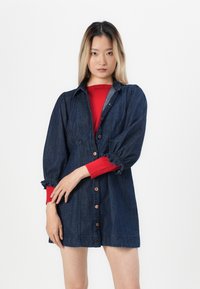 Jeansklänning med knapprad fram, semi-anpassad passform, puffärmar och elastiska ärmslut. Bärs över en röd långärmad topp.