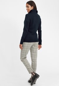 Maglione dolcevita lavorato a maglia di un blu scuro abbinato a pantaloni aderenti a quadri beige e neri. Il modello indossa scarpe nere con suola spessa.