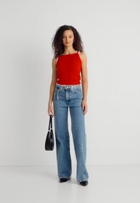 Γυναίκα με σγουρά μαλλιά, ντυμένη με κόκκινη μπλούζα χωρίς μανίκια, τζιν ψηλόμεσο με φαρδιά μπατζάκια, μαύρες ψηλοτάκουνες γόβες, κρατώντας μαύρη τσάντα, στέκεται μπροστά σε απλό φόντο.