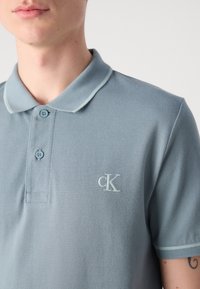 Lys blå polo skjorte med strukturert stoff, to knapper, krage og en liten brodert logo på venstre bryst. Kontrasterende kragekant.