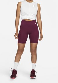 Diep bordeaux hoge shorts en een witte cropped tanktop. Nike-logo op beide. Model draagt witte crew-sokken en bordeaux sneakers met roze accenten.