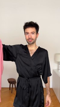 Μαύρο σατένρόμπα με φαρδιά μανίκια και δεμένη μέση, κρατημένη μπροστά σε ροζ ένδυμα. Λείο υλικό, μίνιμαλ σχεδίαση και λειτουργική αισθητική.