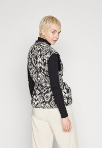 Gilet a righe bianche e nere con un design a peplum, caratterizzato da forme geometriche, indossato sopra una maglietta nera a maniche lunghe e pantaloni color crema.
