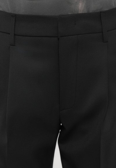Black tailored trousers feature a smooth fabric texture, slim fit, front pleats, and discreet side pockets. Simple waistband design.