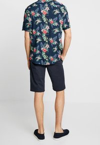 Chemise florale à manches courtes bleu marine avec des motifs rouges et verts, associée à un short sombre. Le mannequin est de dos, portant des chaussures foncées.