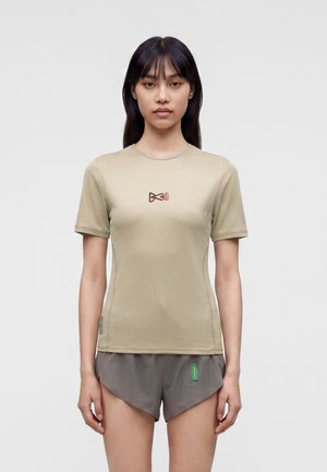 Joven mujer con cabello largo y oscuro lleva una camiseta beige de manga corta con un pequeño logo rojo y negro y pantalones cortos deportivos grises sobre un fondo liso.