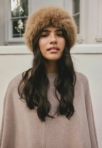 Brown fur hat with a fluffy texture, worn over long dark hair. The individual wears a light brown oversized sweater. Soft fabric, relaxed fit.