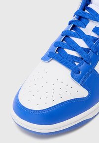 Blå og hvid sneaker med glat læderoverdel, perforeret tåboks, strukturerede blå snørebånd og hvid sål med blå detaljer.