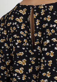 Robe noire à imprimé floral, en tissu texturé avec un motif de fleurs jaunes et blanches. Le dos présente une petite ouverture en forme de goutte fermée par un bouton.