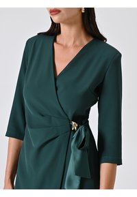 Robe wrap verte en tissu doux, avec un décolleté en V, des manches trois quarts et un accent doré décoratif à la taille.