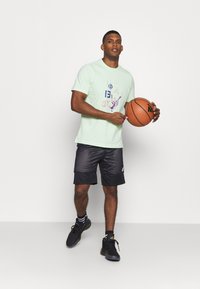 Camiseta verde claro de manga corta con un diseño gráfico multicolor, combinada con pantalones cortos negros hasta la rodilla. Sosteniendo un balón de baloncesto, lleva zapatillas negras.