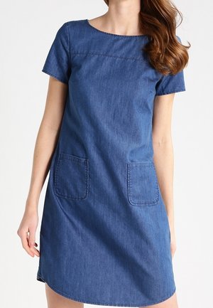 Kobieta w niebieskiej sukience z denimu o krótkich rękawach, długości do kolan, z kwadratowym dekoltem i dwoma naszywanymi kieszeniami z przodu.