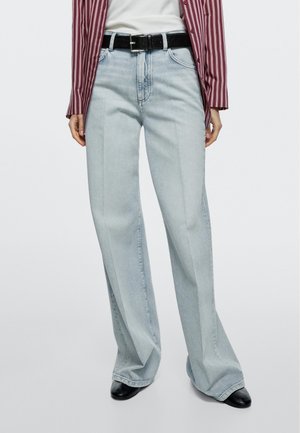 Osoba nosząca jasnoniebieskie dżinsy z szerokimi nogawkami, czarny pasek, biały top oraz koszulę zapinaną na guziki w czerwono-białe paski.