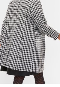 Abrigo de patrón de houndstooth en blanco y negro, con mangas largas, un corte relajado y bolsillos laterales. El dobladillo cuenta con un detalle de piel negra.