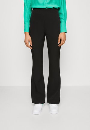 Pantaloni neri a gamba larga con vita alta, realizzati in un tessuto morbido, abbinati a sneakers bianche e una maglietta verde, mani ai fianchi.