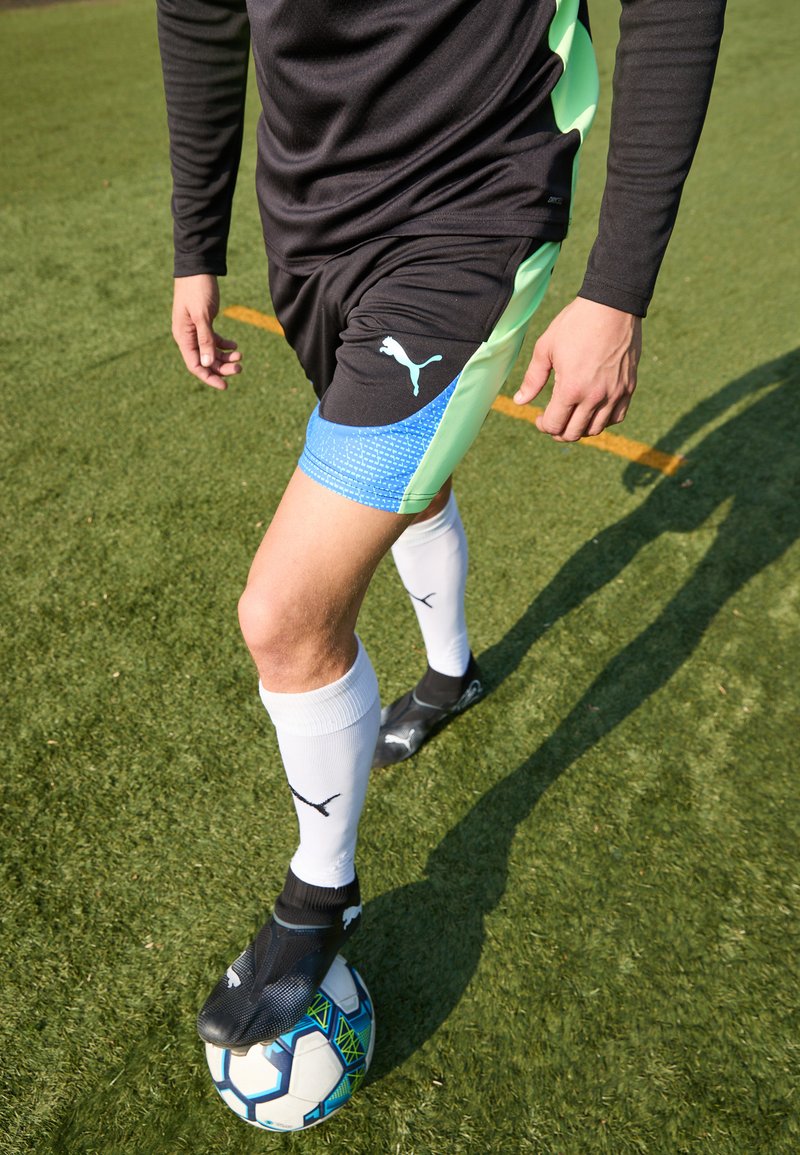 Schwarze Sportshorts mit grünen Akzenten, blau gemustertem Saum und PUMA-Logo; weiße Socken und schwarze Fußballschuhe, auf einem Fußball positioniert.
