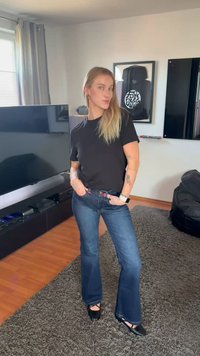 Noor naine, kellel on pikk blondid juuksed, must T-särk, sinised teksapüksid ja mustad kingad, seisab elutoas, kus on televiisor ja tugitoolipatja.