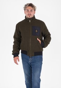 Giacca in pile verde con chiusura a zip, tasca sul petto in blu navy, polsini e orlo a costine, e una vestibilità comoda. Indossata con jeans blu.