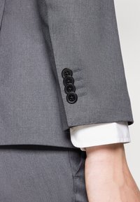 Grijze colbertmouw met vier zwarte knopen en blauwe stikdetails. Witte shirtmanchet zichtbaar eronder. Gelaagd stof met een gladde afwerking.