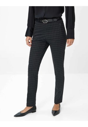 Frau trägt schwarze spitze Ballerinas, dunkle, eng anliegende Hahnentrittmuster-Hosen mit Reißverschlusstaschen, schwarzen Gürtel und schwarze Bluse mit langen Ärmeln.