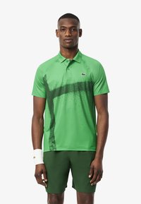 Polo verde con un patrón salpicado y el logo de Lacoste, fabricado en un material ligero; combinado con pantalones cortos verdes a juego.