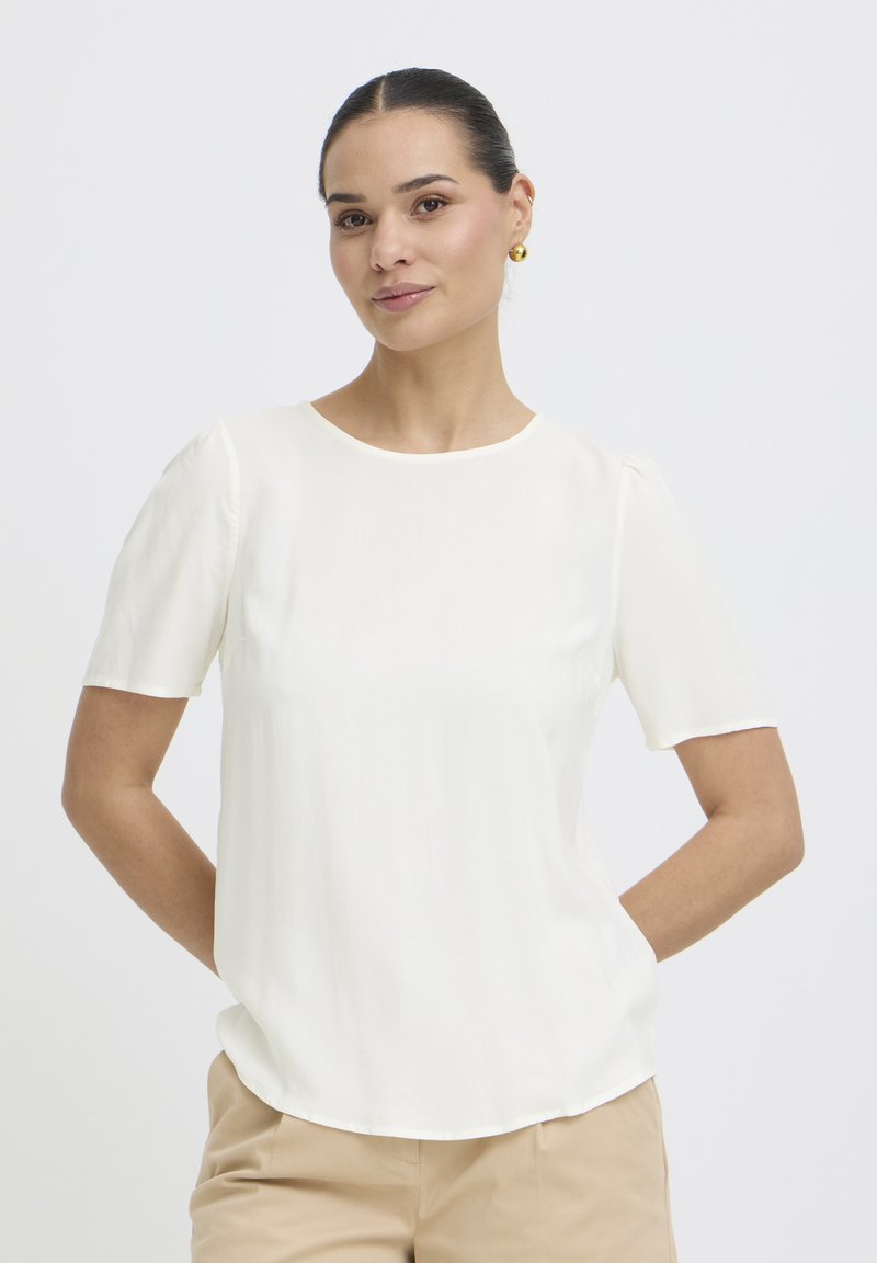 Žena s uhladenými vlasmi, ktorá nosí zlaté náušnice, bielu blúzku s krátkym rukávom a béžové nohavice, ruky za chrbtom, neutrálne pozadie.