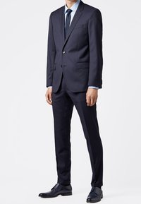 Námořnický oblek na míru se sakem na dva knoflíky, slim-fit kalhotami a světle modrou košilí. Černé společenské boty dotvářejí soupravu.