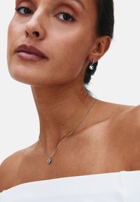 Strieborné náušnice v tvare srdca a zodpovedajúci prívesok na retiazke zdobia modelku. Dizajn je elegantný s lesklým povrchom, na bielom pozadí.