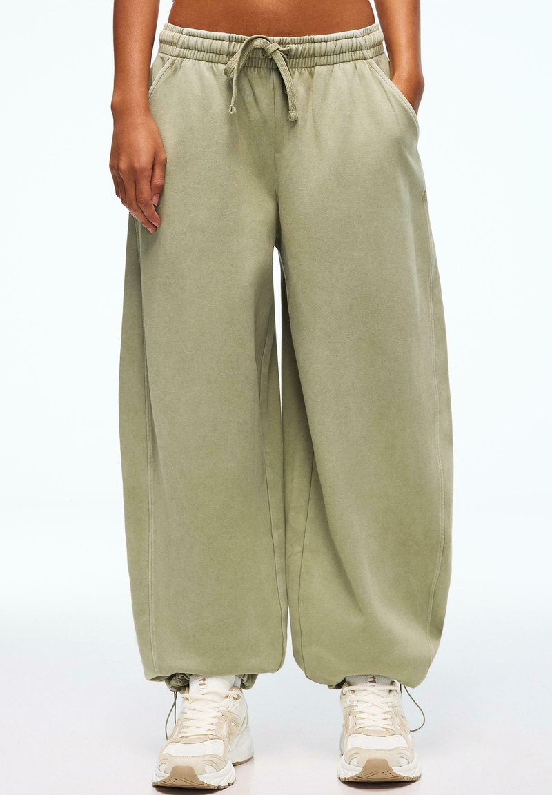 Personne portant un pantalon de survêtement ample vert clair à cordon de serrage avec des poignets élastiques et des baskets blanches épaisses, les mains dans les poches.