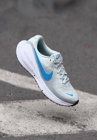 Běžecká obuv Nike s lehkým šedým síťovaným svrškem a modrými akcenty, s polstrovanou podrážkou a zaobleným designem špičky.