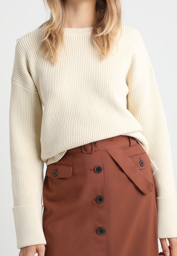 Pull en maille côtelée crème à manches longues associé à une jupe marron avec un design portefeuille diagonal et des accents de boutons noirs.
