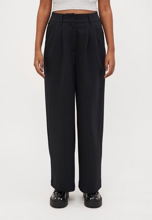 Czarne spodnie o szerokich nogawkach i wysokim stanie z plisami z przodu, noszone z czarnymi ćwiekowanymi butami na platformie oraz krótkim szarym topem.