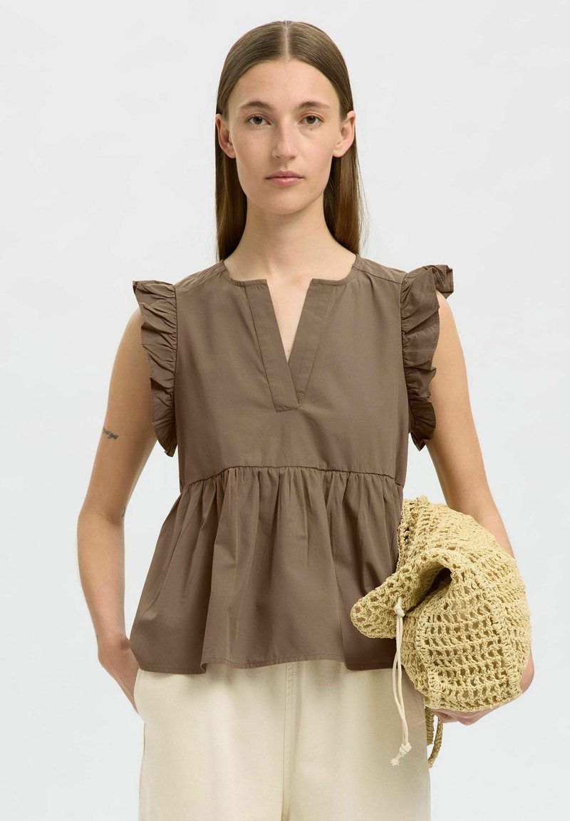 Brązowy top peplum z dekoltem w serek i falbaniastymi rękawami. Wykonany z lekkiego materiału, z marszczoną talią i gładką fakturą. Modelka trzyma siatkową torbę.
