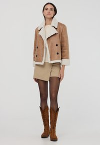 Giacca di shearling marrone con un colletto e polsini in pelliccia sintetica bianca, abbinata a una gonna beige e stivali da cowboy marroni al ginocchio.