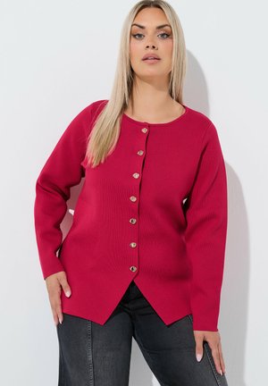 Donna con lunghi capelli biondi che indossa un cardigan rosa scuro con bottoni e jeans scuri, in piedi su sfondo bianco.