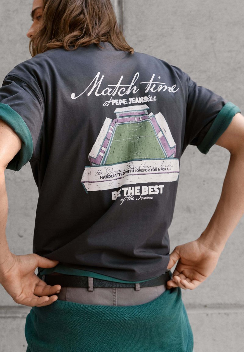 Άνδρας ρυθμίζει τη μέση του, φοράει σκούρο T-shirt με γραφικό ενός ποδοσφαιρικού σταδίου και το κείμενο "Match time, Be the best of the season."