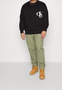 Svart sweatshirt med "CK"-logotyp, gröna cargobyxor och beige arbetsskor med snören. Avslappnad outfit med en ledig passform.