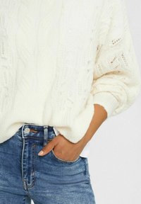 Dikke witte gebreide trui met een kabelontwerp, met verlaagde schouders en geribde manchetten, gecombineerd met donkerblauwe high-waisted jeans.