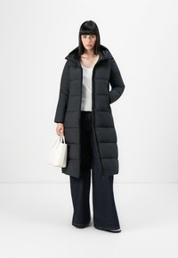 Zwarte pufferjas met capuchon, ritssluiting en gestikte ontwerp; gecombineerd met een witte top en wijde jeans. Model houdt een kleine witte handtas vast.