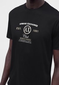 T-shirt noir Armani Exchange porté par une personne à la peau foncée, avec un texte blanc et beige et des logos centrés sur la poitrine.