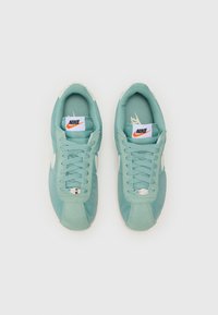Nike sneakers in light teal featuring suede and mesh materials, white accents, lace-up closure, and branded tongue tags.