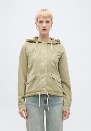 Osoba s krátkými blond vlasy, oblečená v světle olivové kapuci se šňůrkami a světle modrých džínách, čelící dopředu na jednobarevném pozadí.