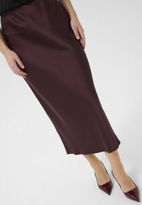 Burgundy A-linje kjol i slätt tyg, som når mitt på vadbenet, parat med glansiga spetsiga klackskor. Strukturerad yta synlig.