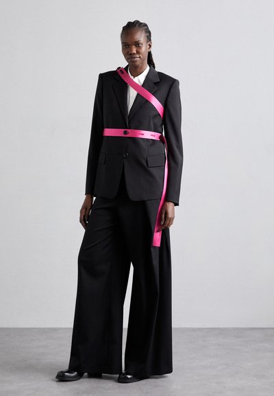 Costume noir ajusté avec des jambes larges et un accent en ruban rose. La veste présente un seul bouton et des poches, assortie à une chemise blanche.